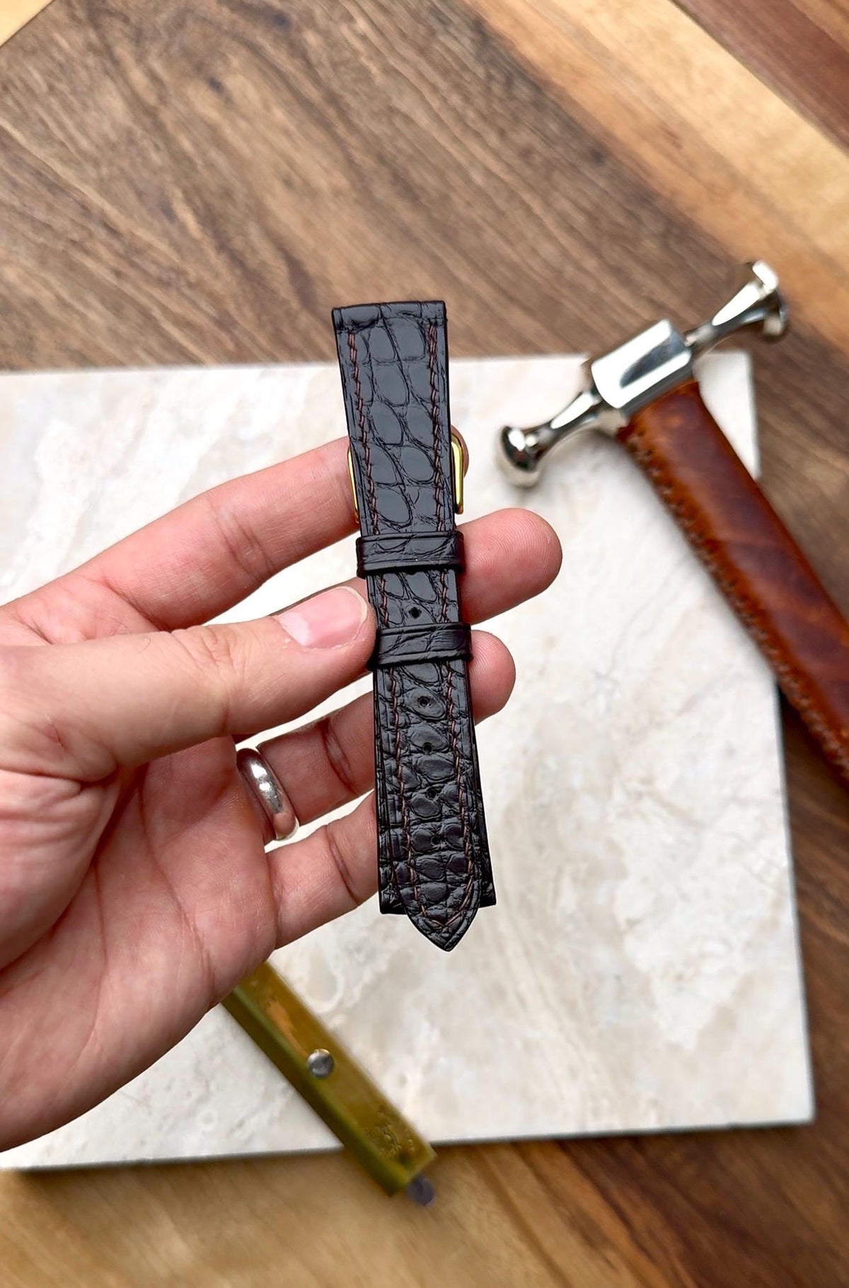 Handmade dark alligator watch strap held above the workbench, with a hammer and tools on a marble block